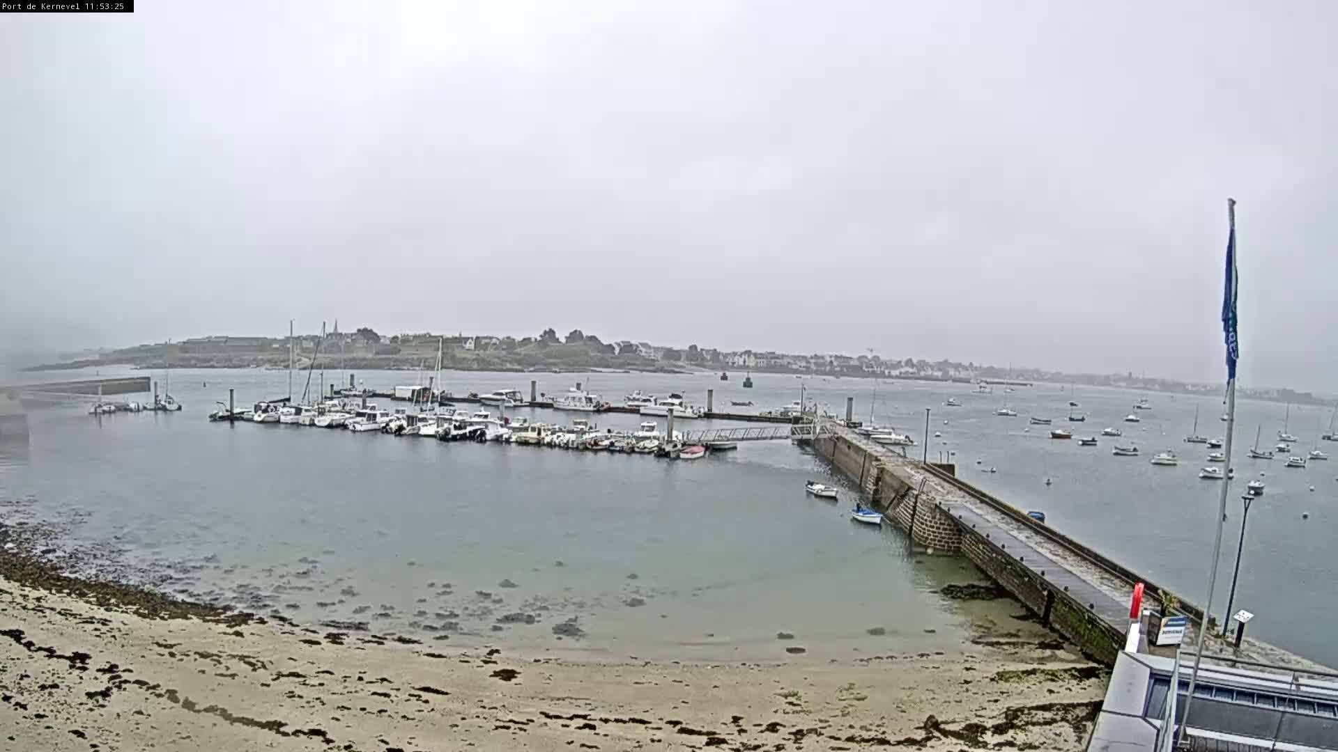A harbor filled with numerous boats is seen on an overcast day, with a sandy beach and a stone pier in the foreground.