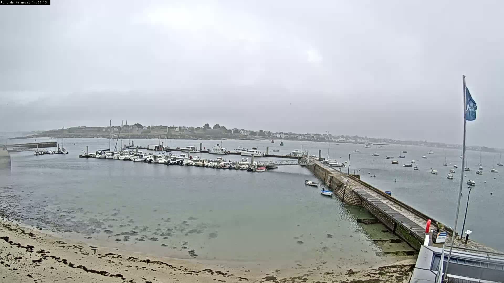 A harbor filled with numerous boats is seen on an overcast day, with a sandy beach and a stone jetty in the foreground.