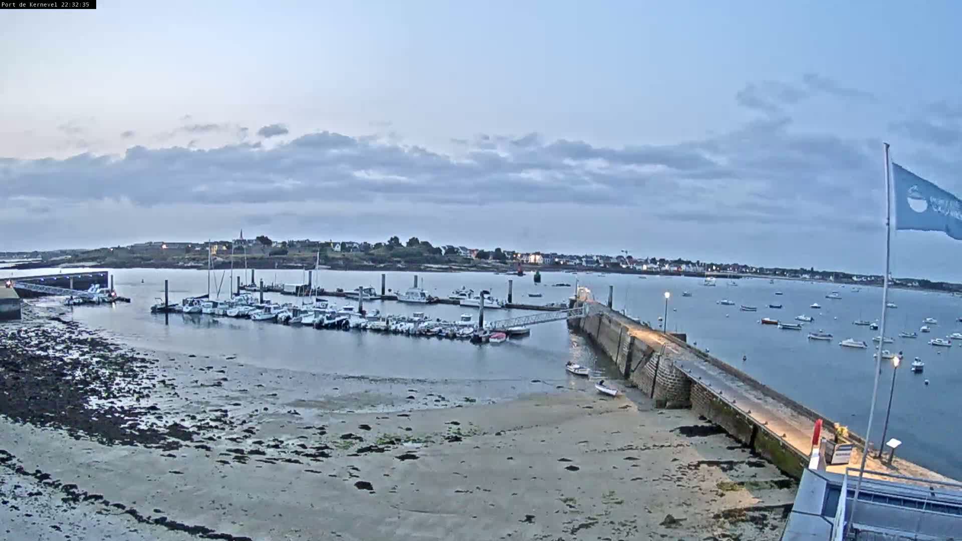 A calm, overcast evening scene shows a marina filled with boats, a long pier extending into the water, and a sandy shore at low tide.