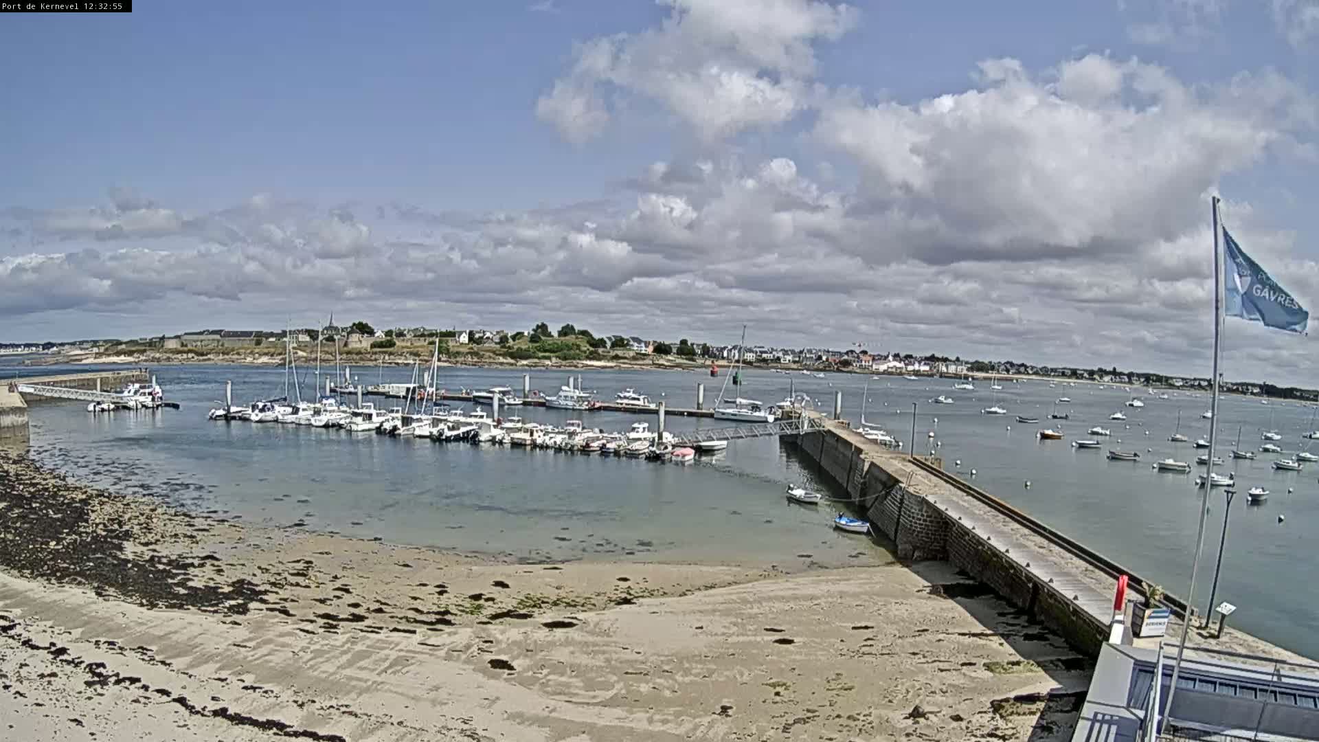 A harbor filled with numerous boats is visible under a partly cloudy sky, with a sandy shoreline in the foreground.