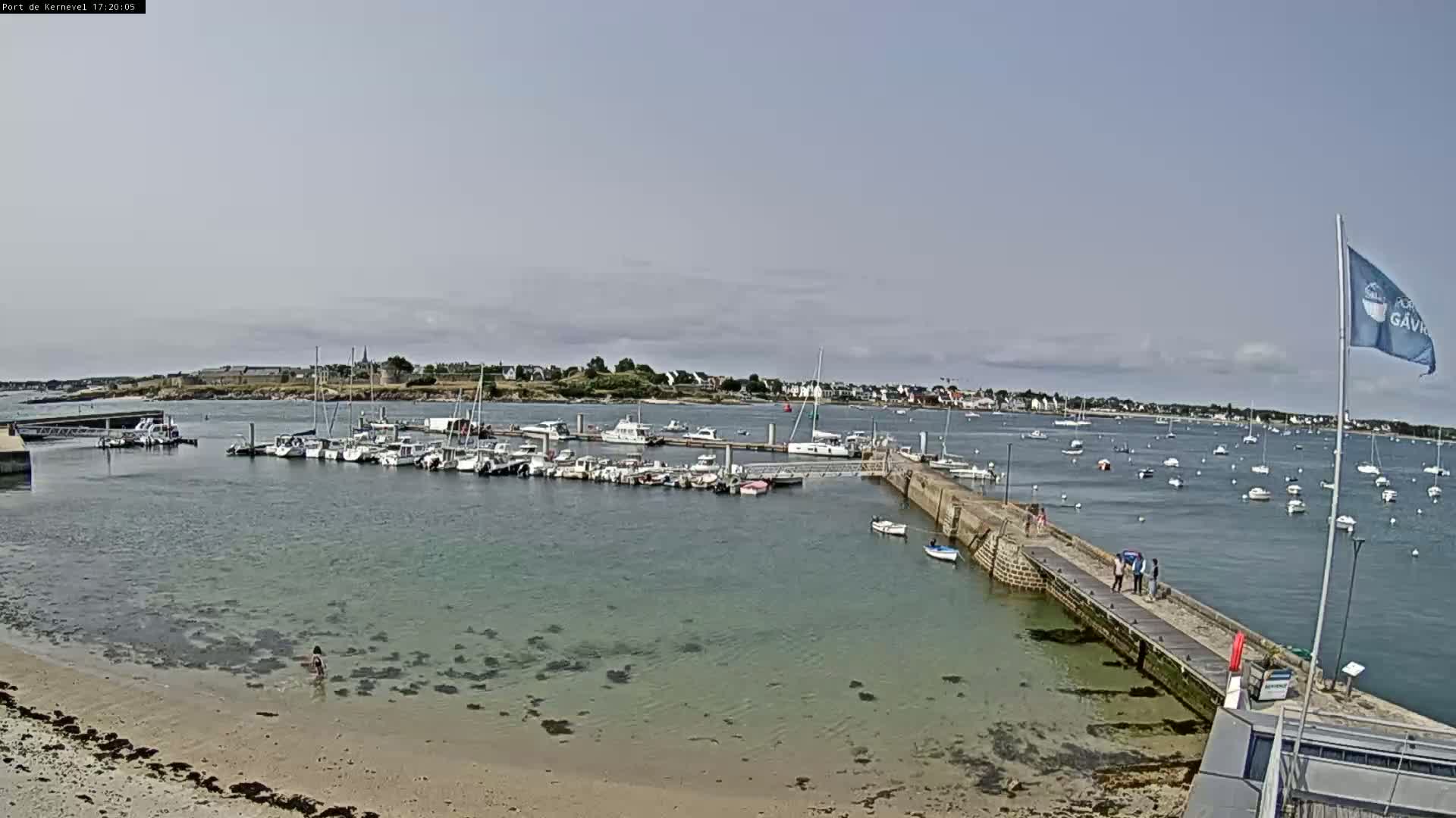 A marina filled with boats is situated on a calm, sunny day next to a sandy beach and a small town in the distance.