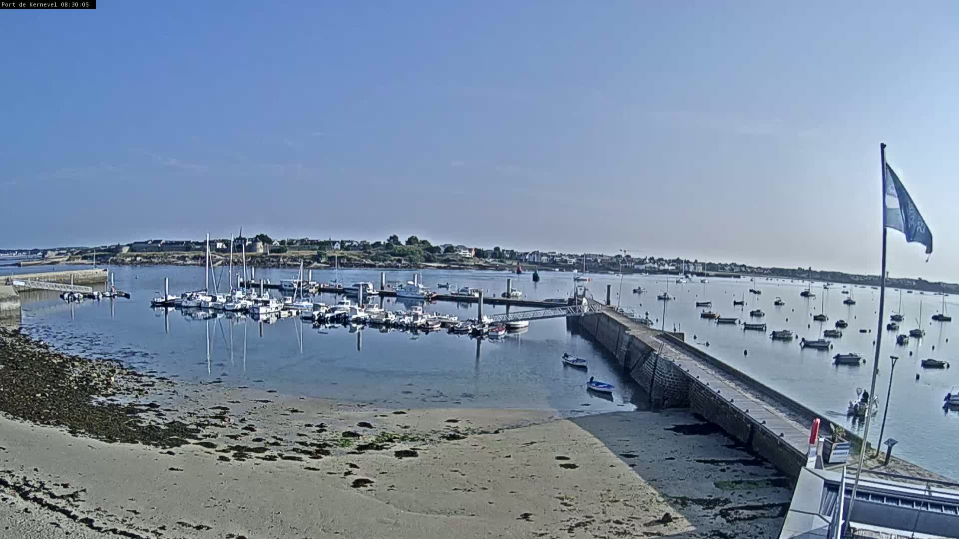 A calm, sunny day shows a harbor filled with numerous boats and yachts, a sandy shore, and a small town in the distance.