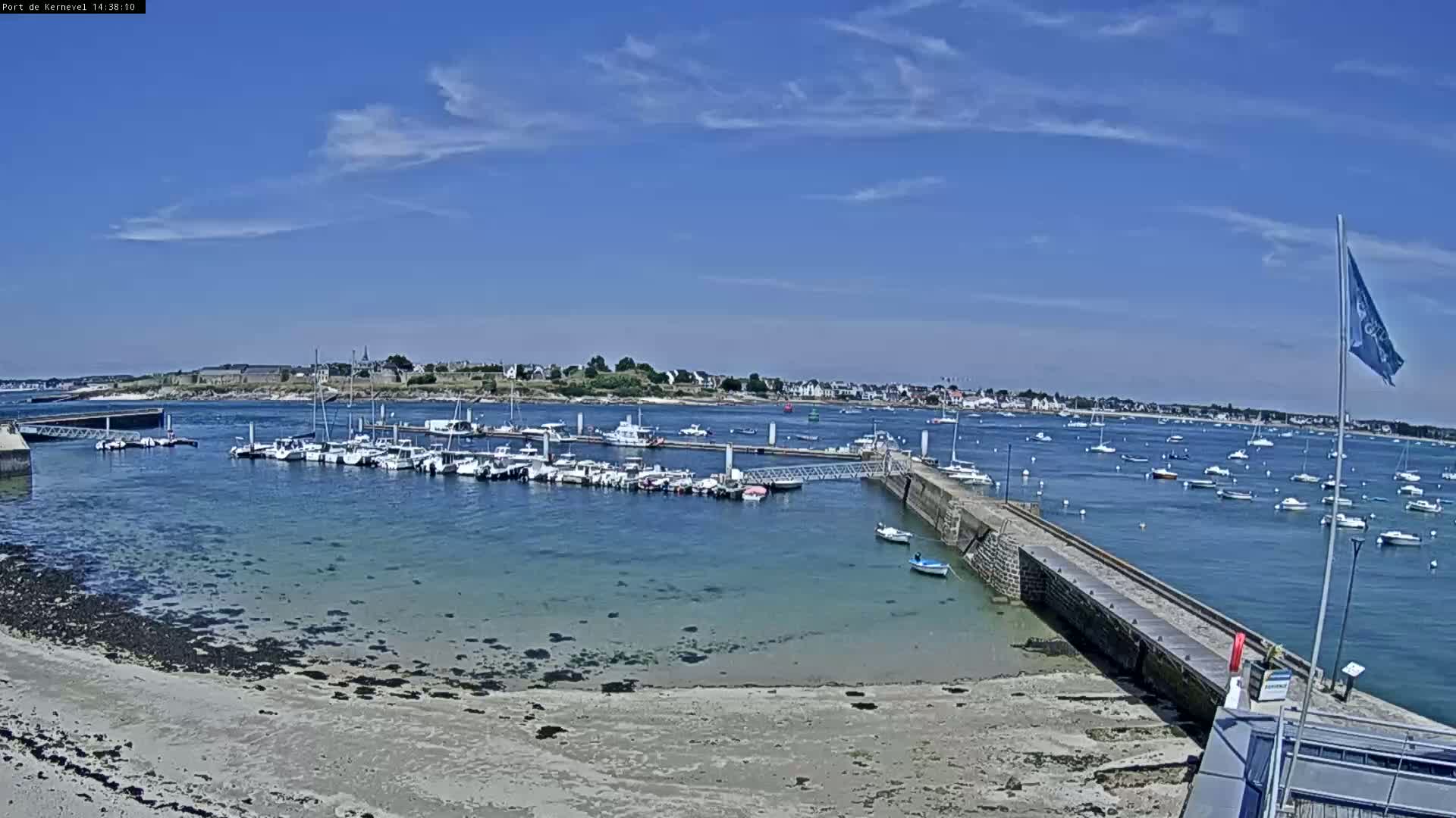 A marina filled with numerous boats is situated on a calm, sunny day next to a sandy beach and a stone breakwater, with a town visible in the distance across the water.