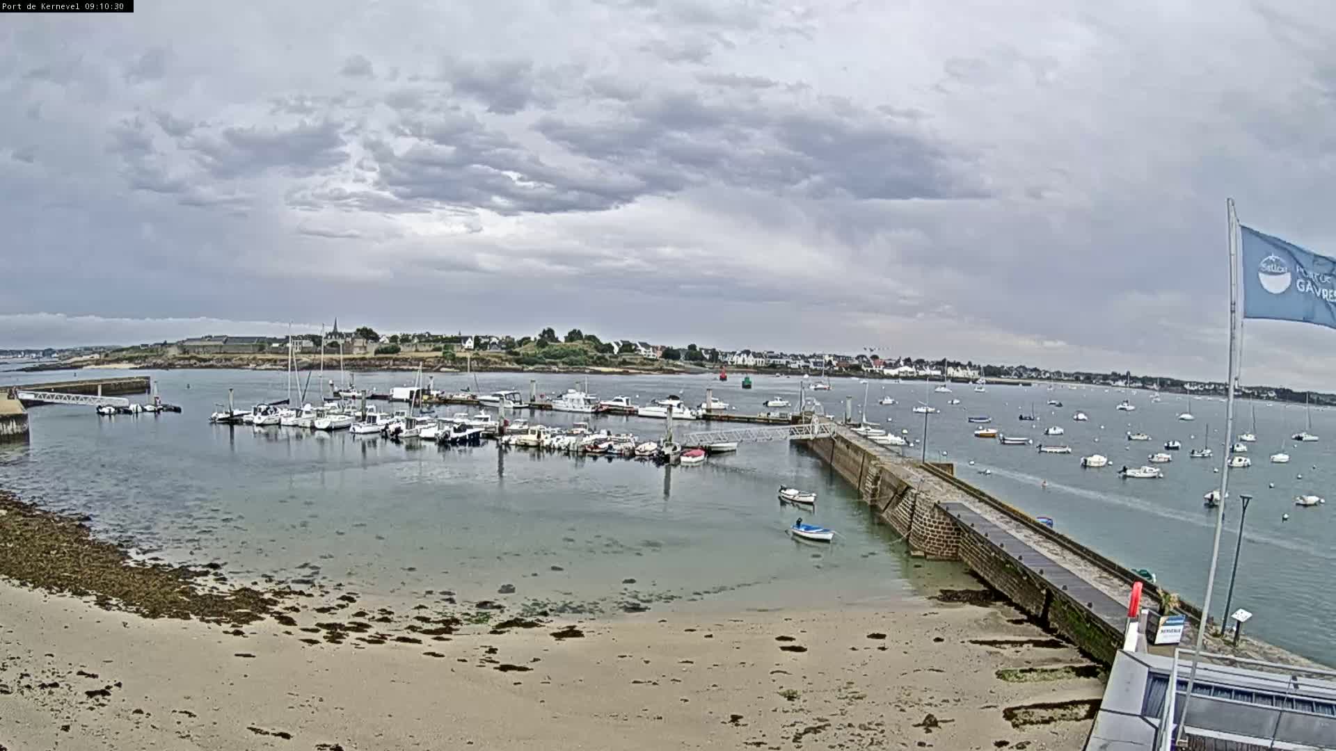 A harbor filled with numerous boats is situated on a calm body of water under an overcast sky, with a sandy beach visible in the foreground.