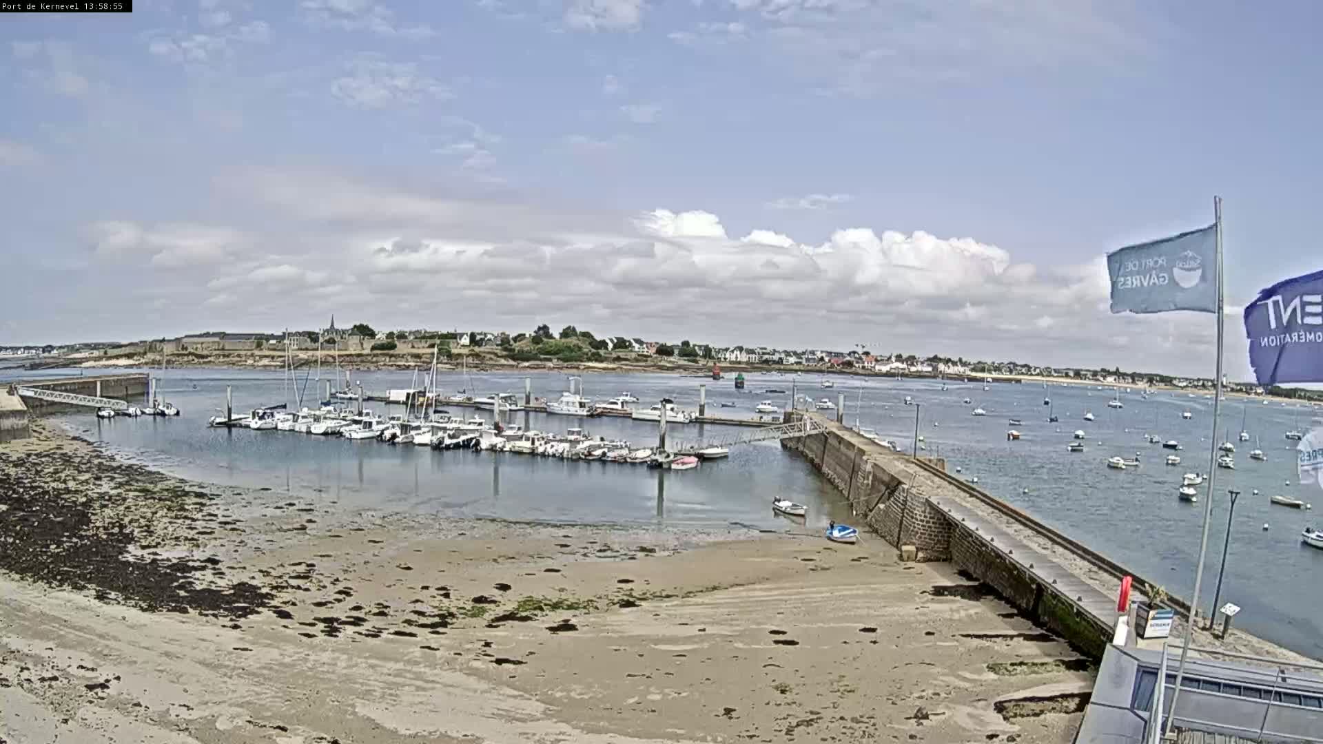 A harbor filled with numerous boats is seen under a partly cloudy sky, with a sandy shoreline in the foreground and a town visible in the distance.