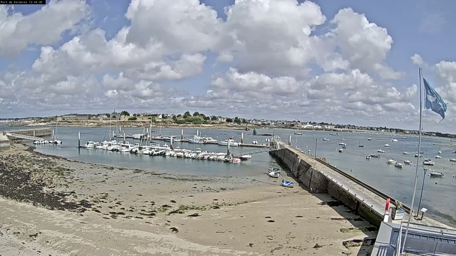 A harbor filled with numerous boats sits under a partly cloudy sky, with a sandy shore visible in the foreground.