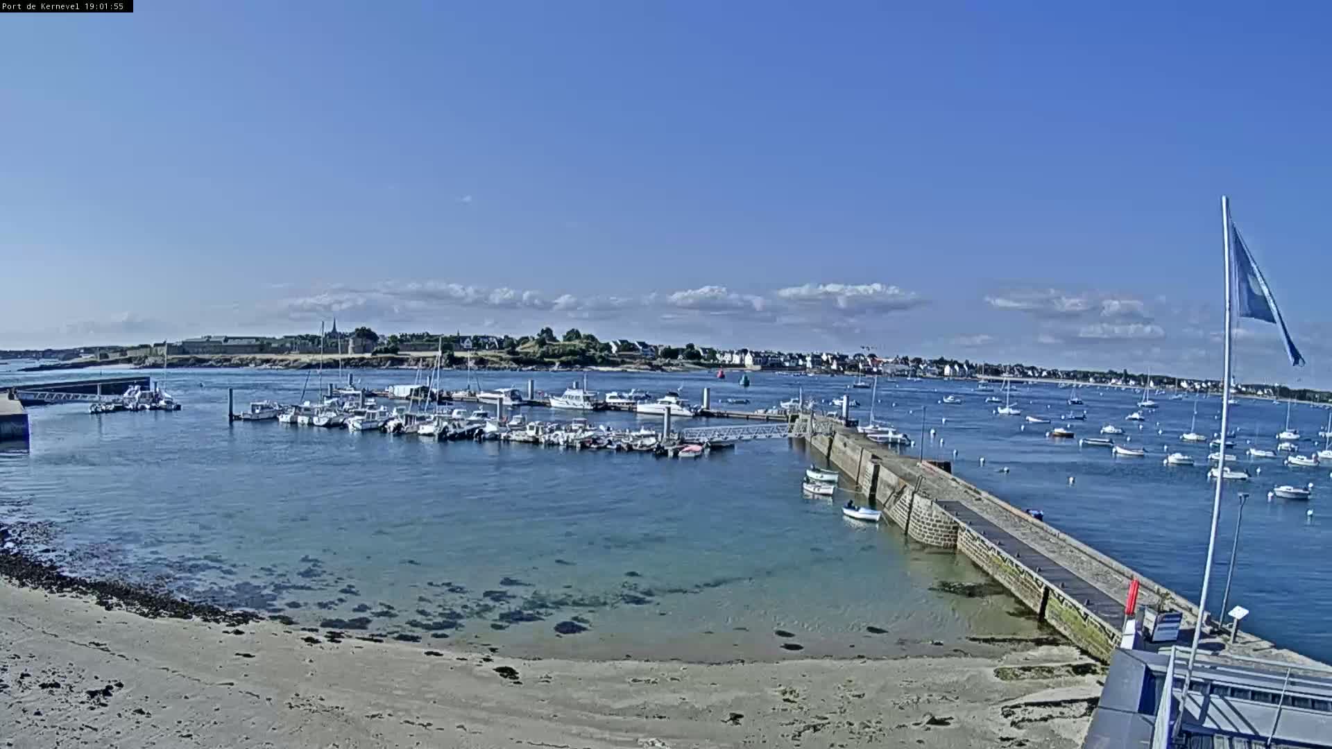 A marina filled with numerous boats is situated near a sandy beach under a mostly sunny sky with some scattered clouds.