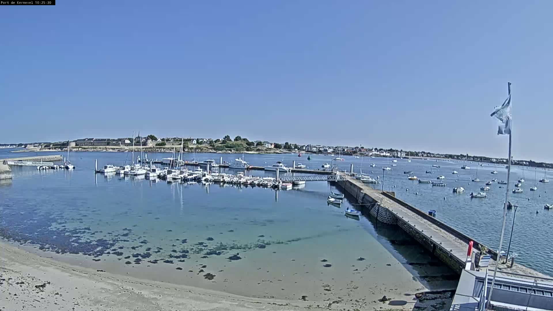 A sunny day shows a marina filled with sailboats and motorboats, a sandy beach, and calm, clear water extending to a town in the distance.