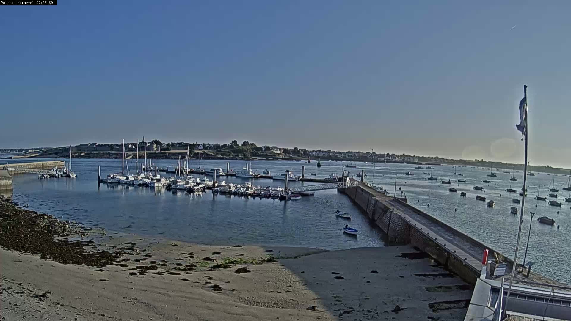 A calm harbor filled with numerous boats is seen on a sunny day, with a sandy beach and a stone jetty in the foreground.