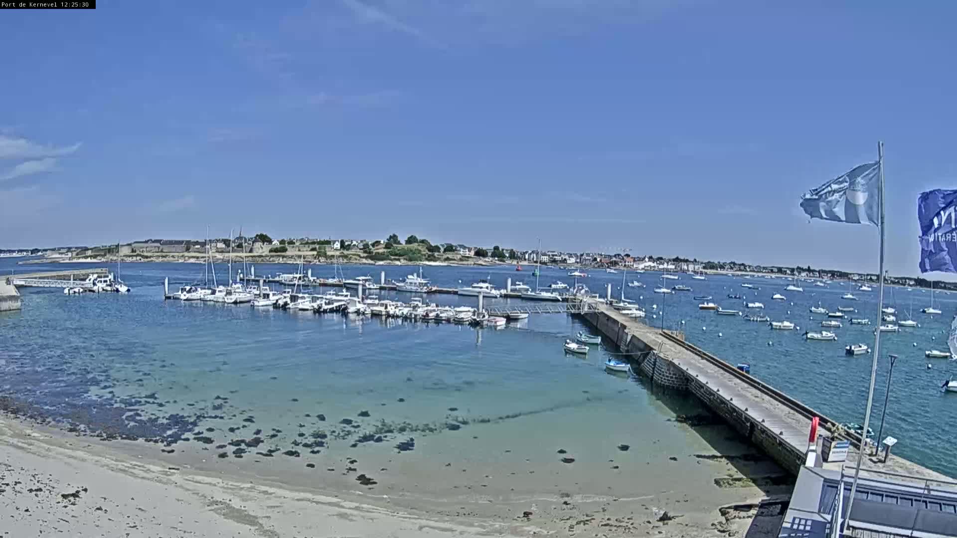 A sunny day shows a harbor filled with boats, a sandy beach, and a town in the distance.