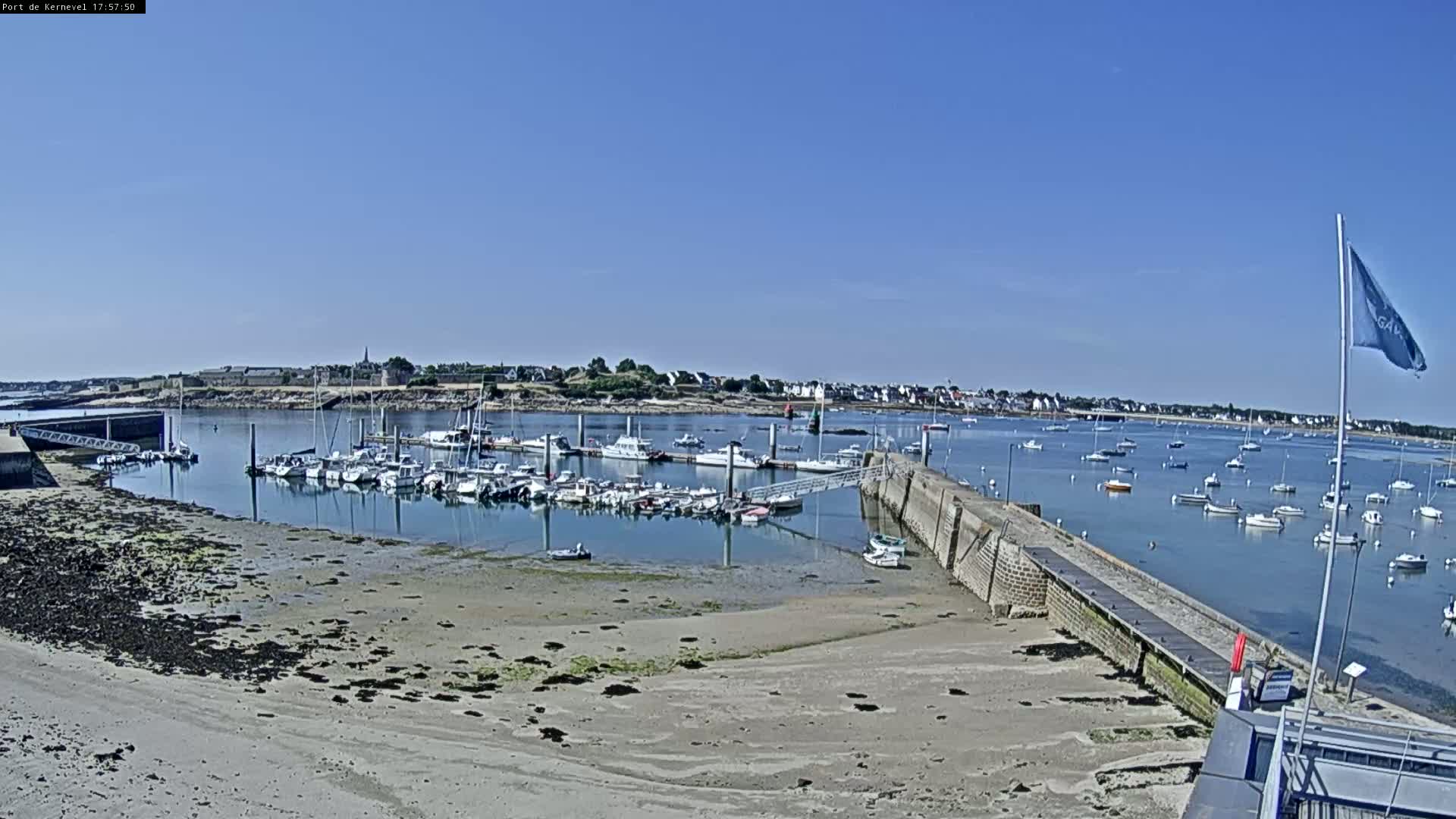A harbor filled with numerous boats is situated on a calm, sunny day next to a sandy shore and a stone pier.