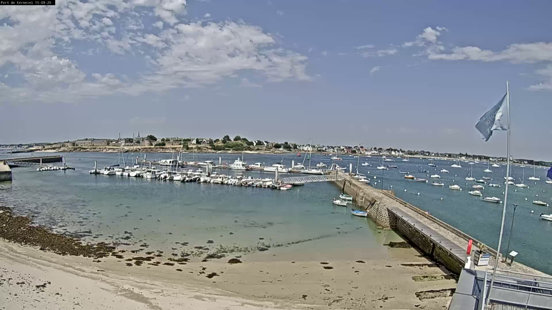 A sandy beach and calm harbor filled with numerous boats are visible under a partly cloudy sky.