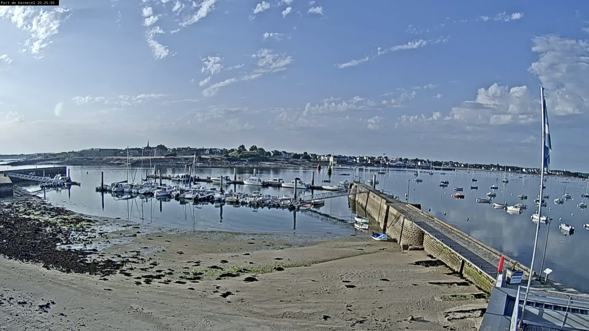 A mostly sunny day shows a harbor filled with many boats, docked and afloat, with a sandy shore and a stone walkway extending into the water.