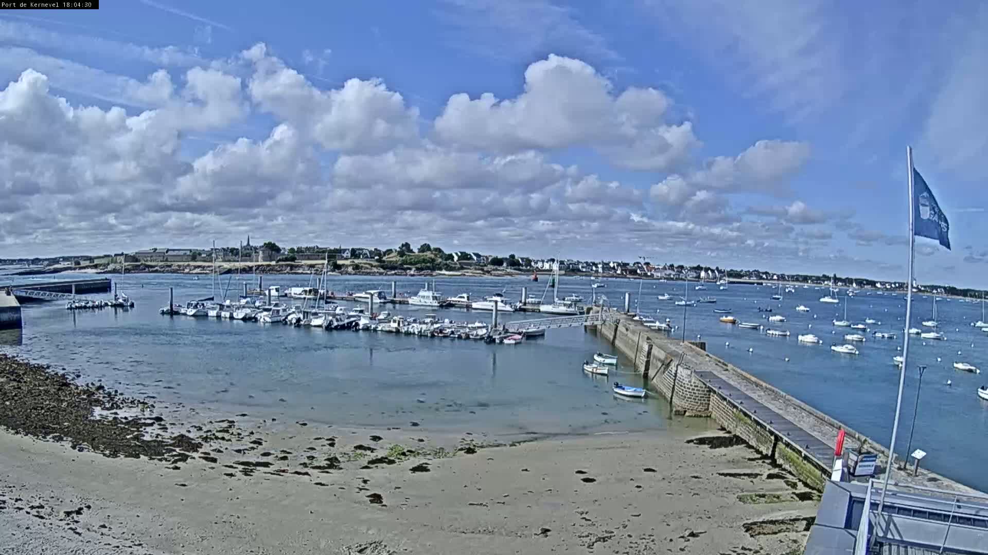 A mostly sunny day shows a harbor filled with numerous boats, a sandy shore, and a town in the distance.