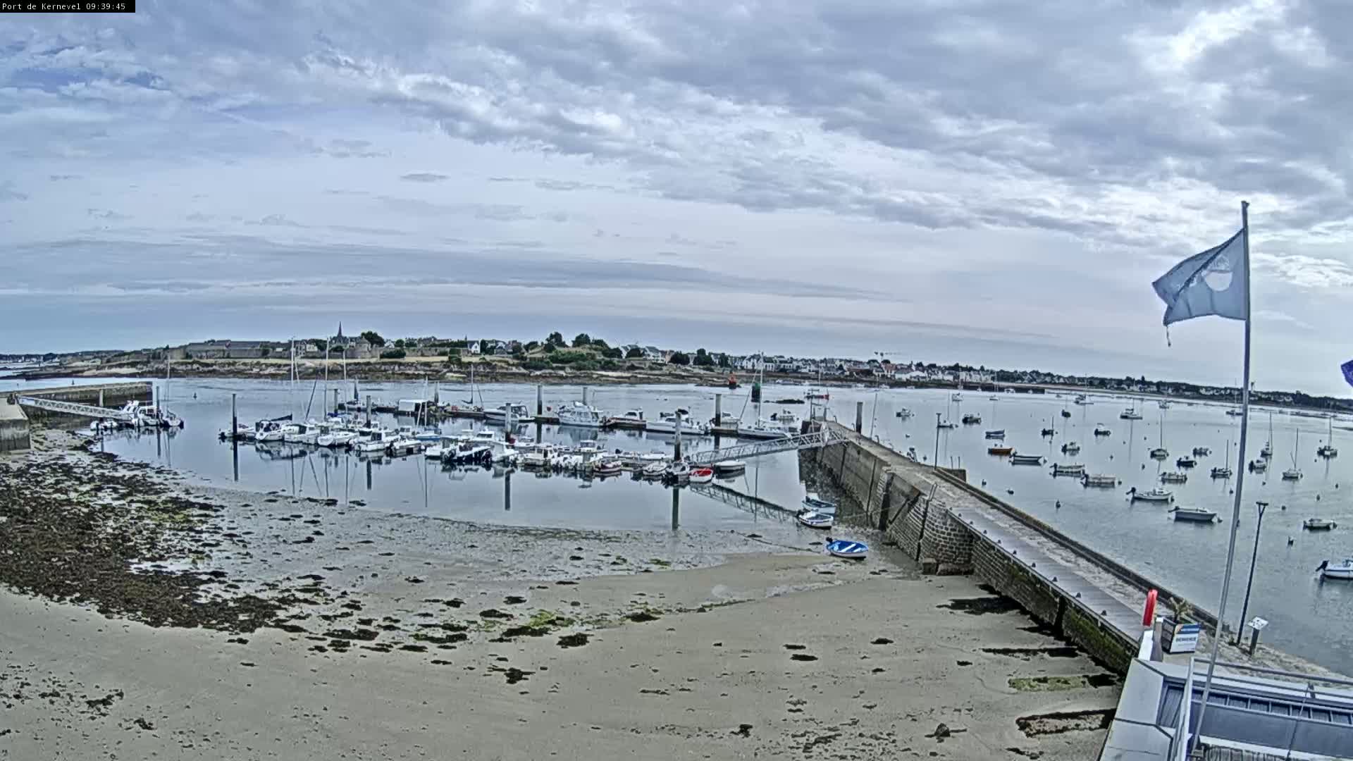 A harbor filled with numerous boats sits under a cloudy sky, with a sandy shore visible in the foreground.