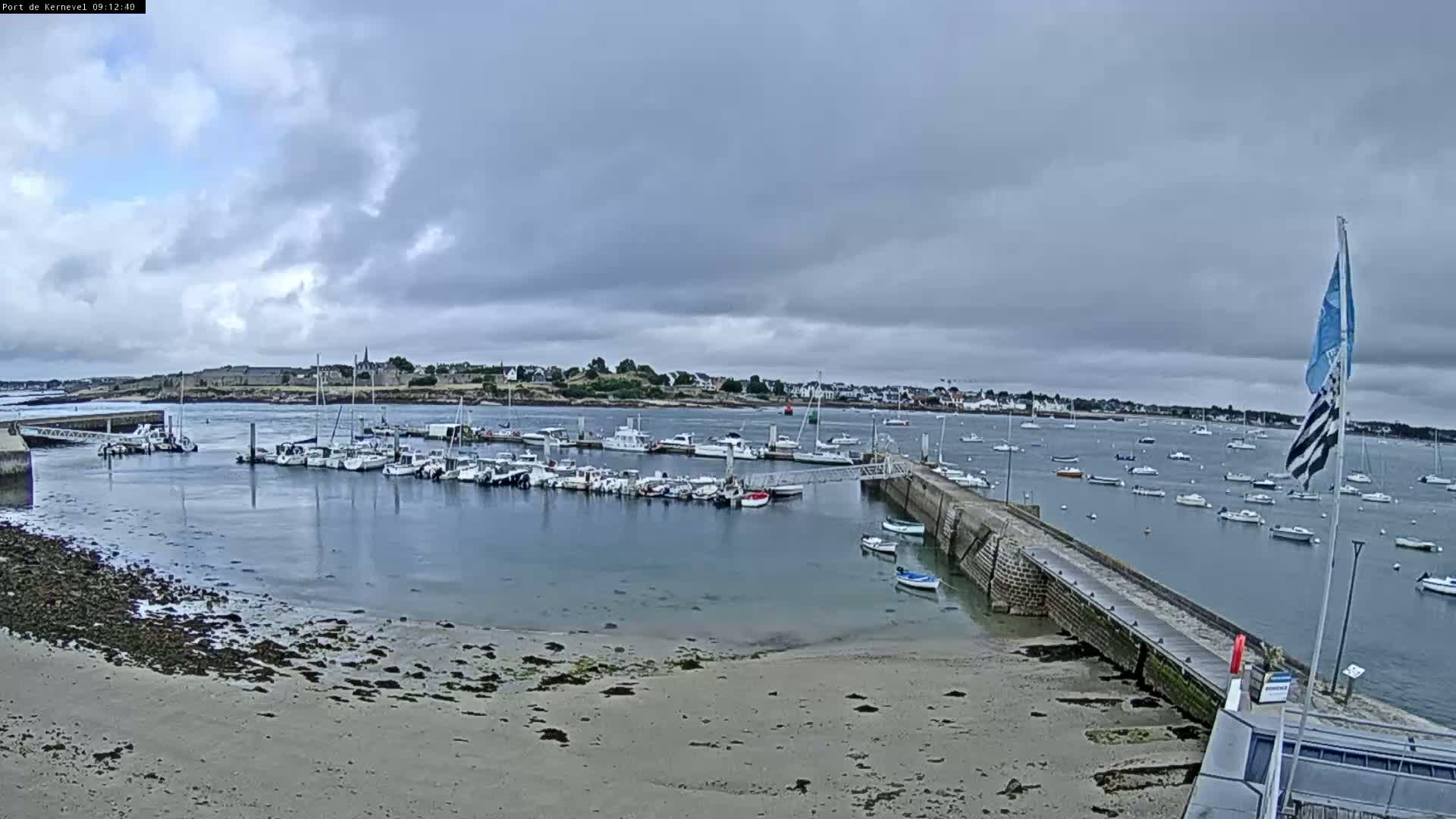 A harbor filled with numerous boats is seen under a cloudy sky, with a sandy beach and a stone pier in the foreground.