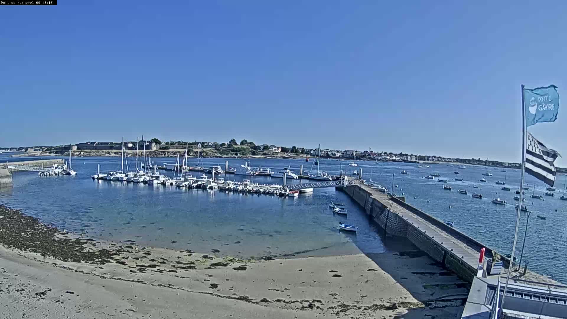 A harbor filled with numerous boats is situated on a calm, sunny day next to a sandy beach and a stone jetty.