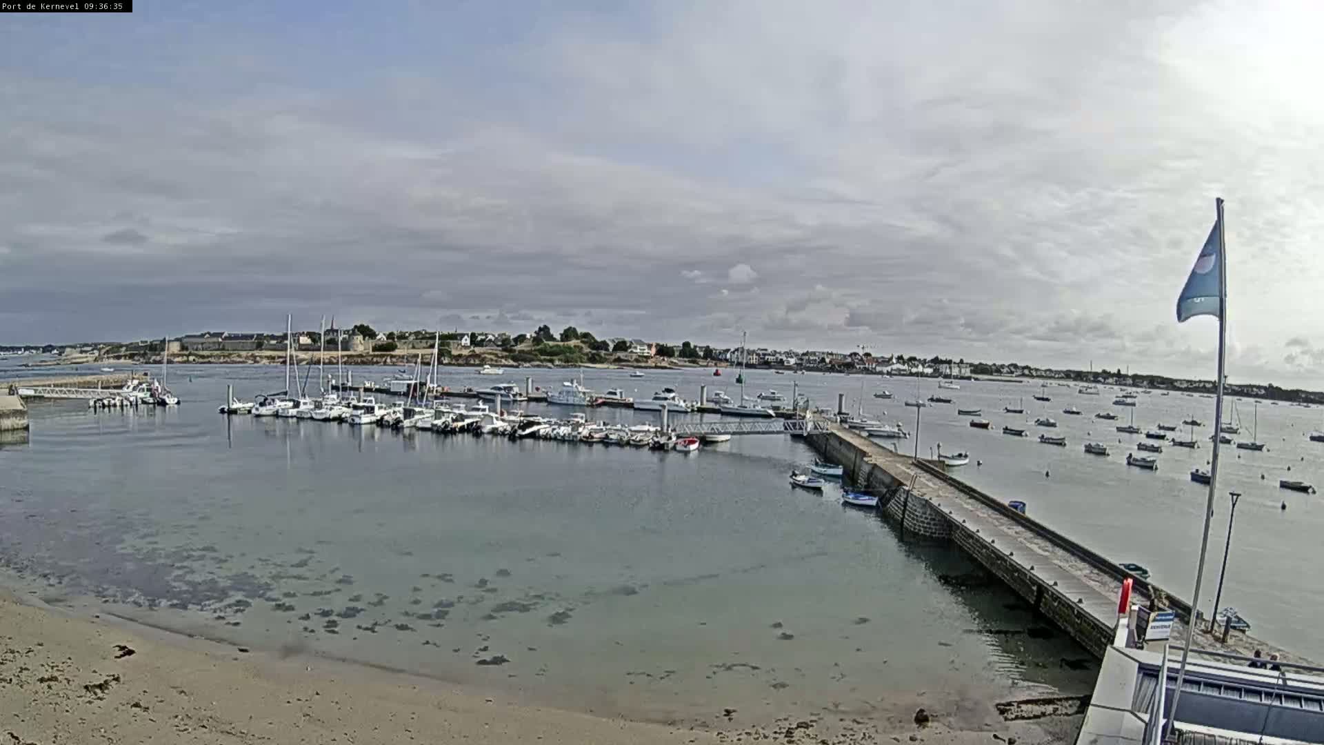 A marina filled with numerous boats is situated on a calm body of water under a cloudy sky, with a sandy beach visible in the foreground.