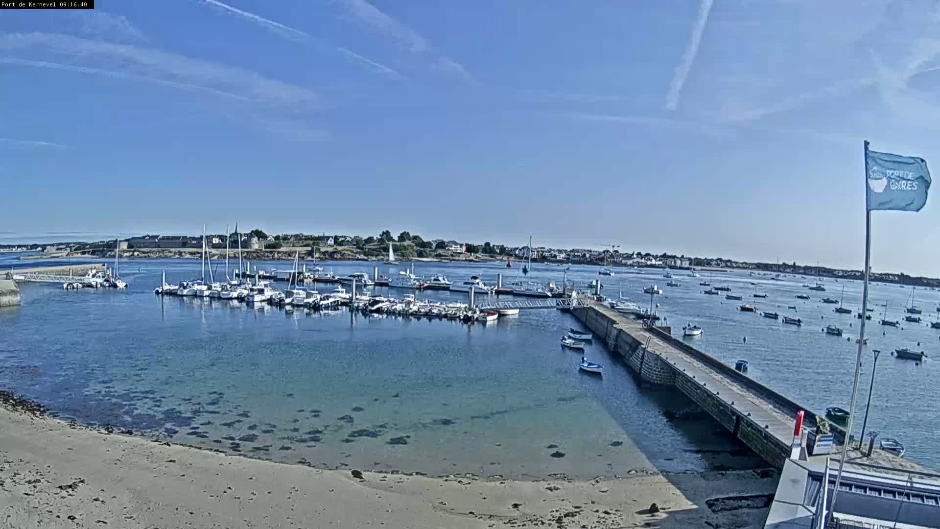 A harbor filled with numerous boats is seen on a sunny day, with calm, clear water and a sandy shore in the foreground.