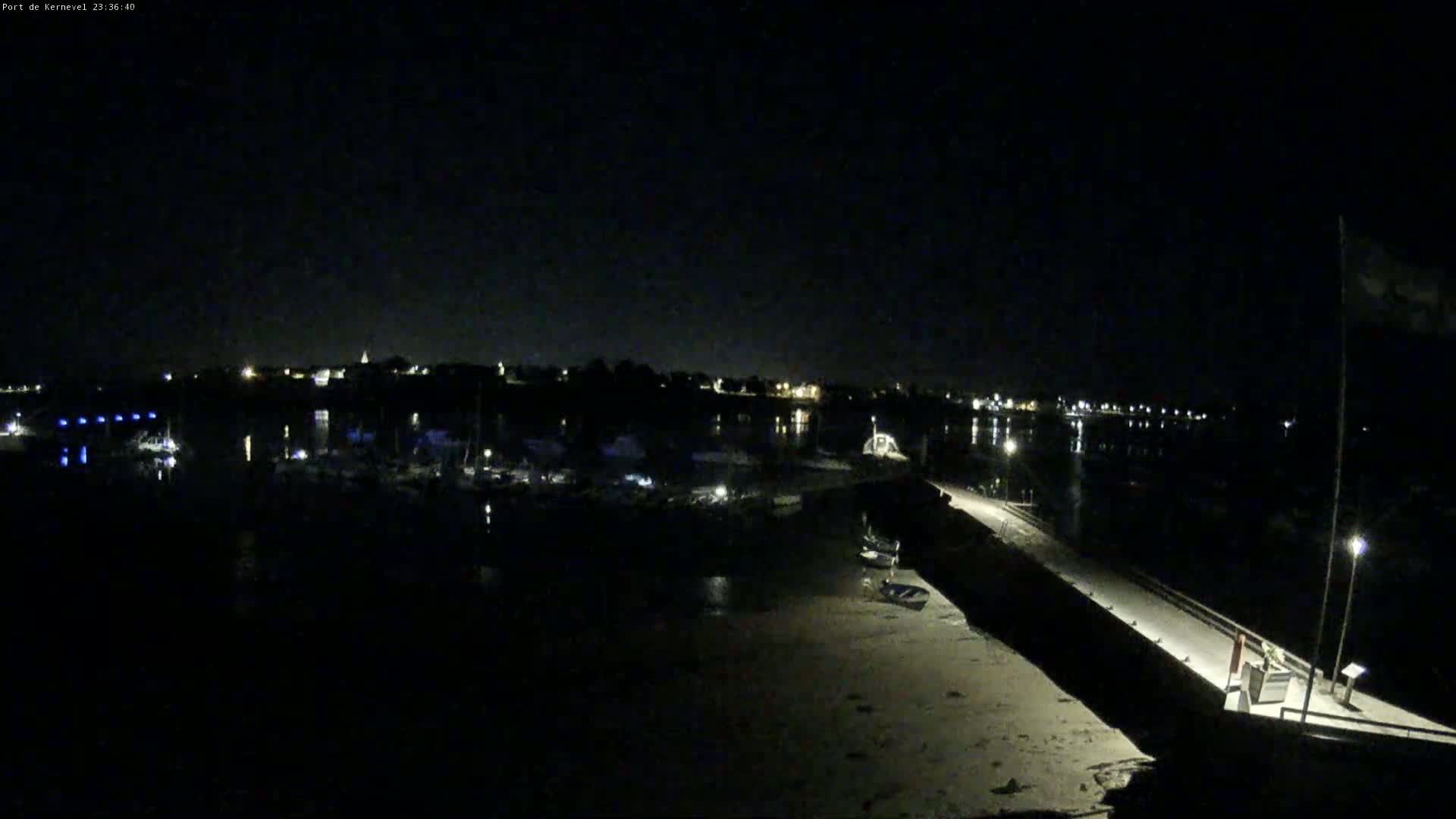 A nighttime view of a harbor with boats moored, a long pier extending into the water, and a town illuminated in the distance under a clear, dark sky.