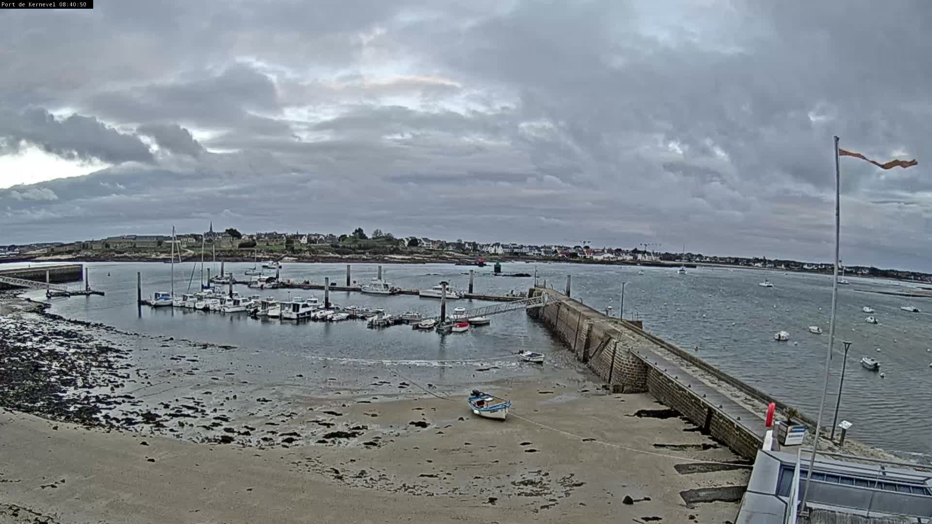 On an overcast day, a coastal harbor is seen with many small boats docked at pontoons and anchored in the grey water, a stone pier extending into the sea, a small boat resting on the exposed sandy shore, and a town lining the distant coast under heavy grey clouds.