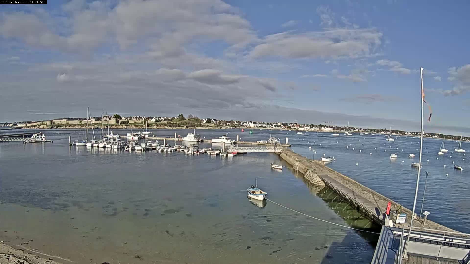 On an overcast day, a coastal harbor is seen with many small boats docked at pontoons and anchored in the grey water, a stone pier extending into the sea, a small boat resting on the exposed sandy shore, and a town lining the distant coast under heavy grey clouds.