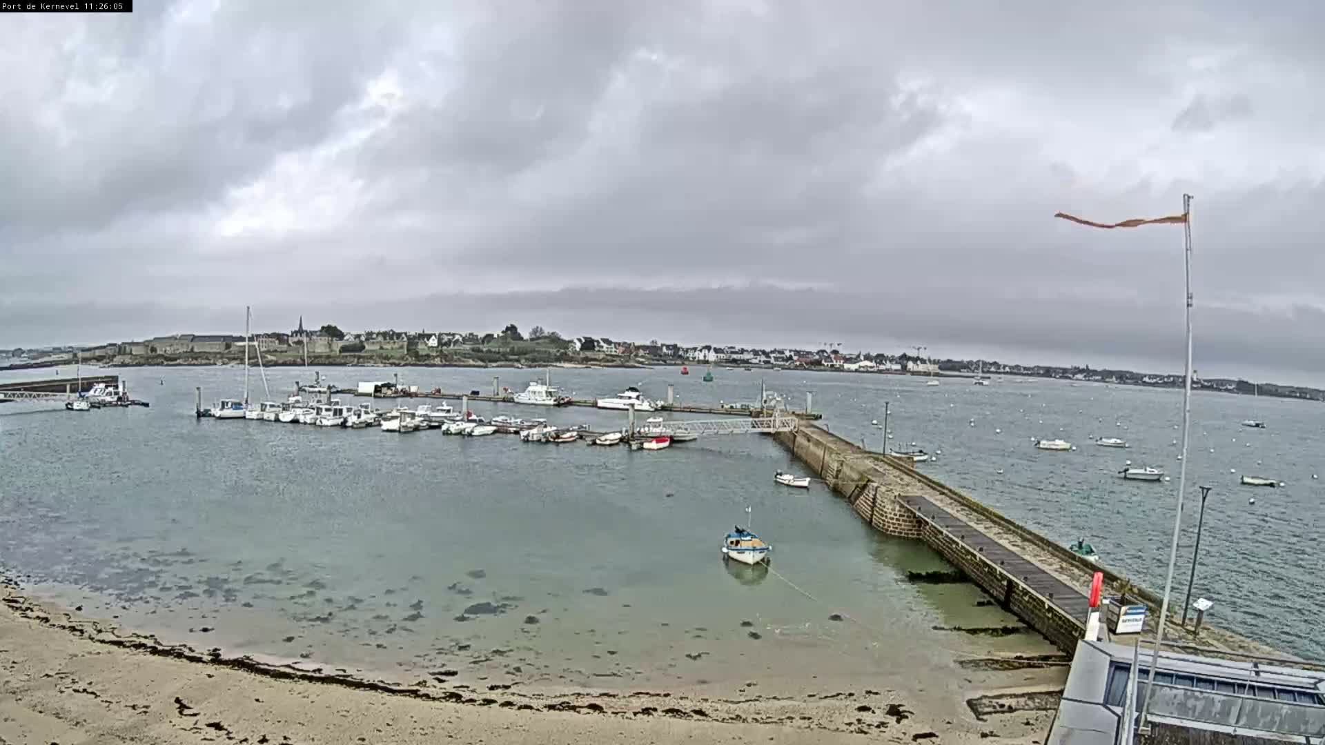 On an overcast day, a coastal harbor is seen with many small boats docked at pontoons and anchored in the grey water, a stone pier extending into the sea, a small boat resting on the exposed sandy shore, and a town lining the distant coast under heavy grey clouds.