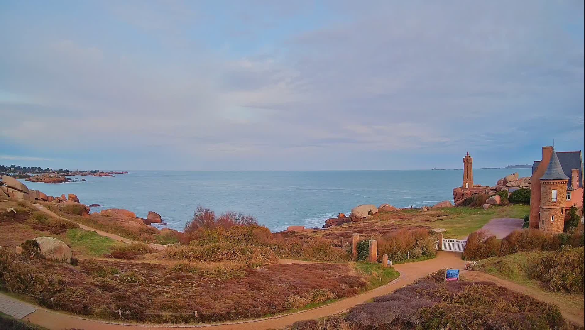 A coastal path winds through low-lying vegetation toward a stone lighthouse and chateau overlooking a calm sea under a cloudy sky.