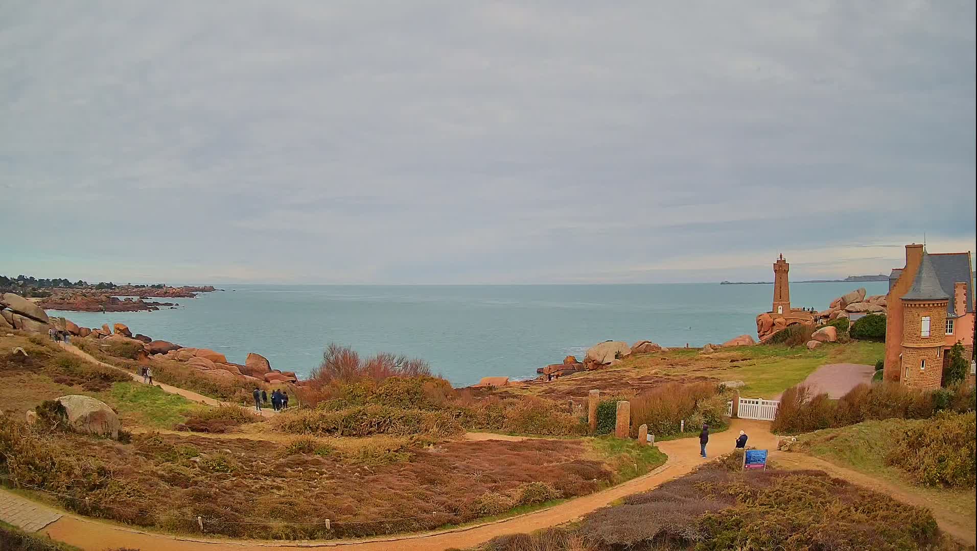 A coastal scene under an overcast sky shows a rocky shoreline with a lighthouse and a stone building, winding paths, and a few people walking.