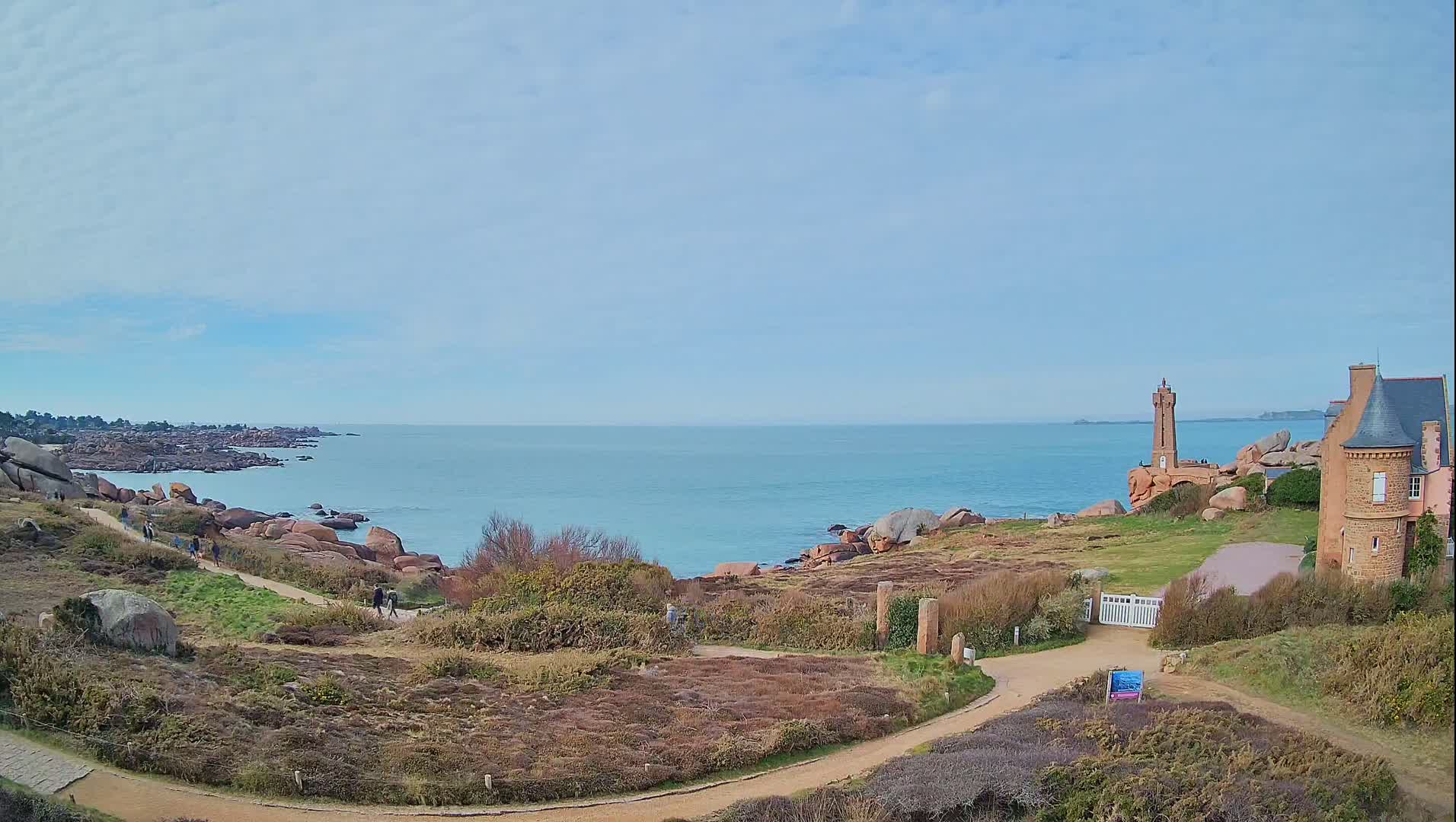 A coastal path winds through low-lying vegetation toward a lighthouse and a stone building overlooking a calm sea under a partly cloudy sky.