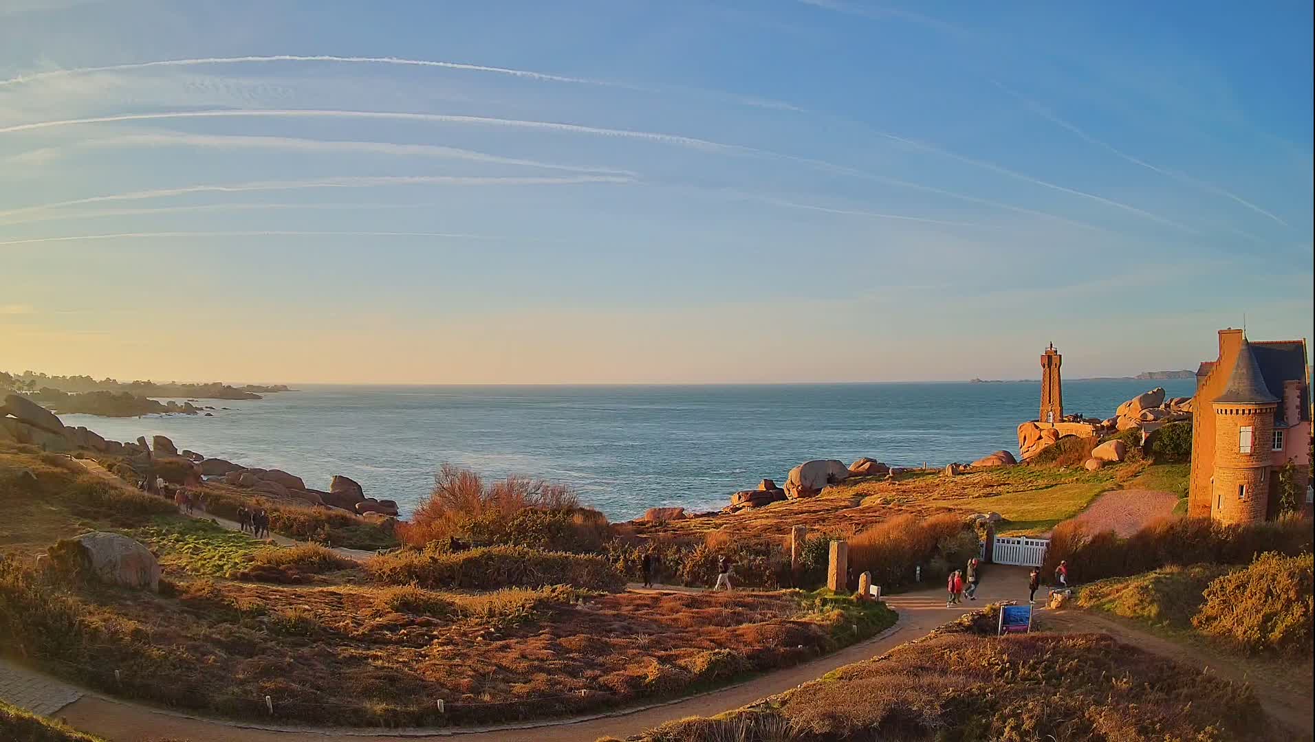 A coastal scene at sunset shows a stone building, a lighthouse, and people walking along a path overlooking a calm ocean under a clear, blue sky with contrails.