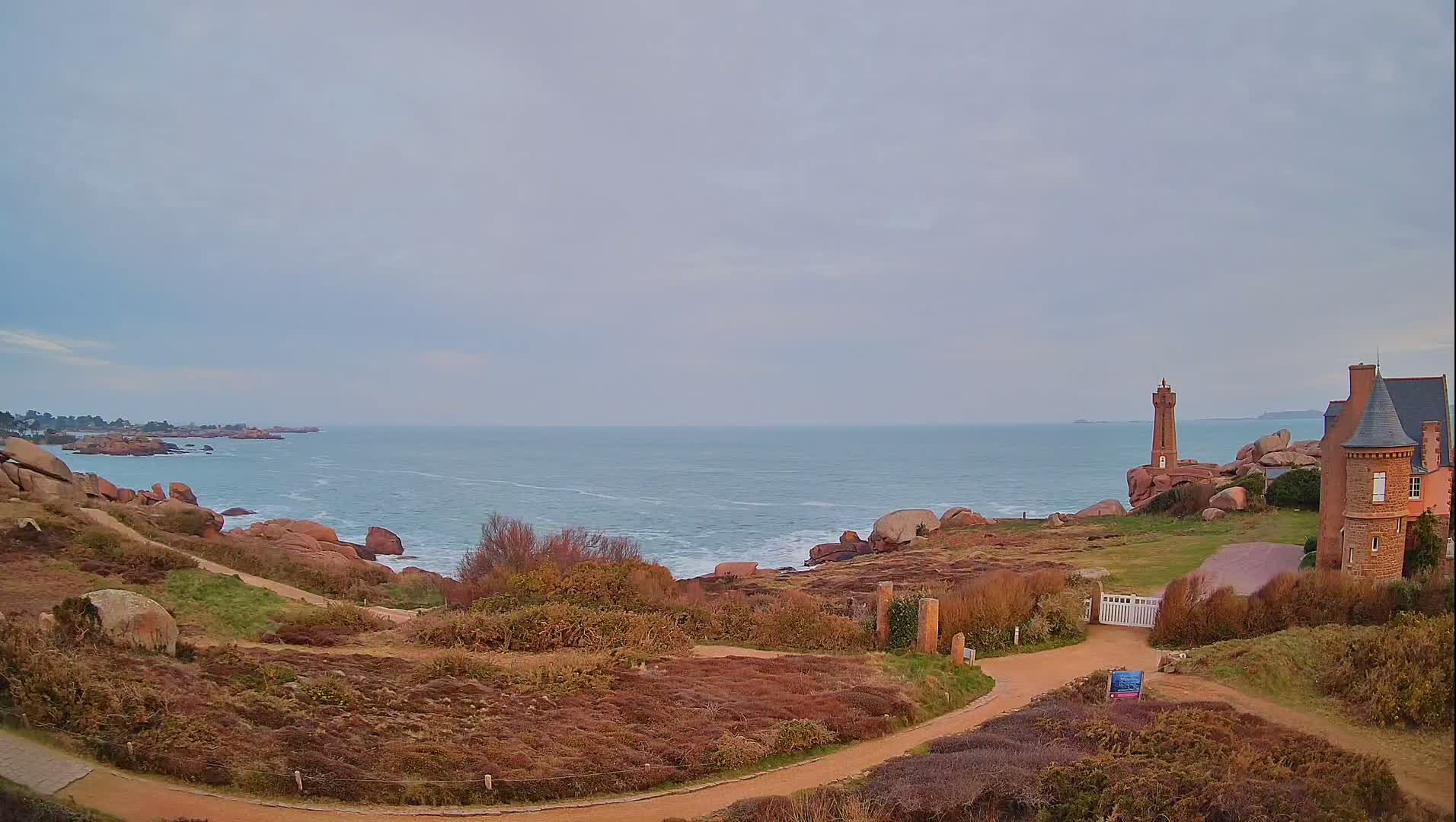 A coastal scene under a cloudy sky shows a stone path winding through low-lying vegetation towards a gated entrance to a stone house and a small lighthouse perched on pink rocks overlooking a calm ocean.