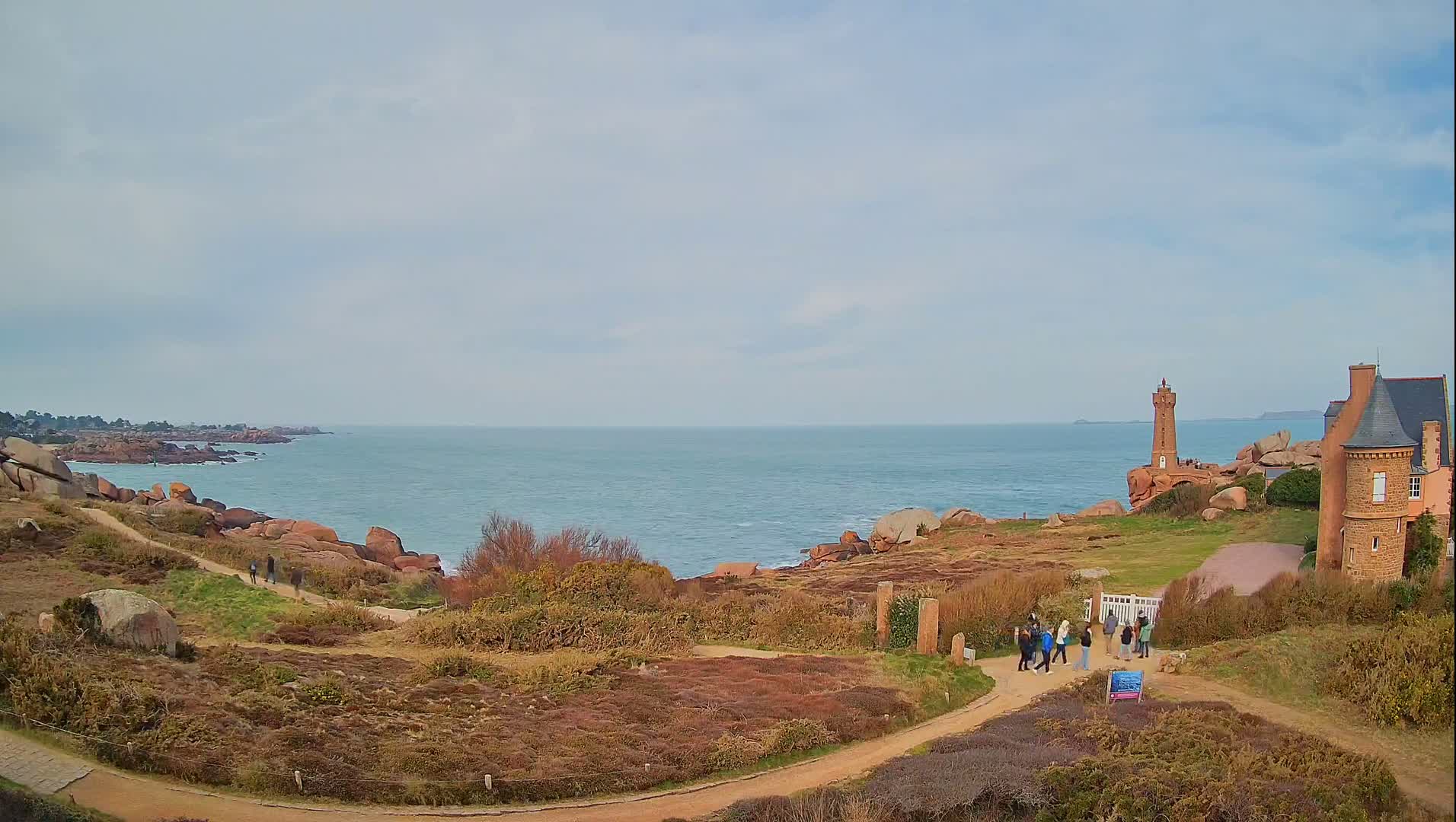 A coastal scene with a lighthouse and stone building overlooking a calm, blue ocean under a cloudy sky.