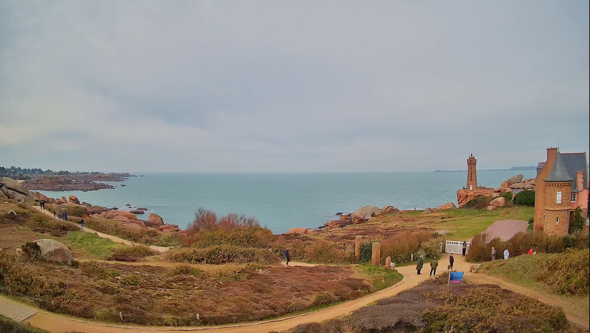 A coastal path winds through low-lying vegetation toward a lighthouse and a stone building overlooking a calm, light-blue sea under an overcast sky.