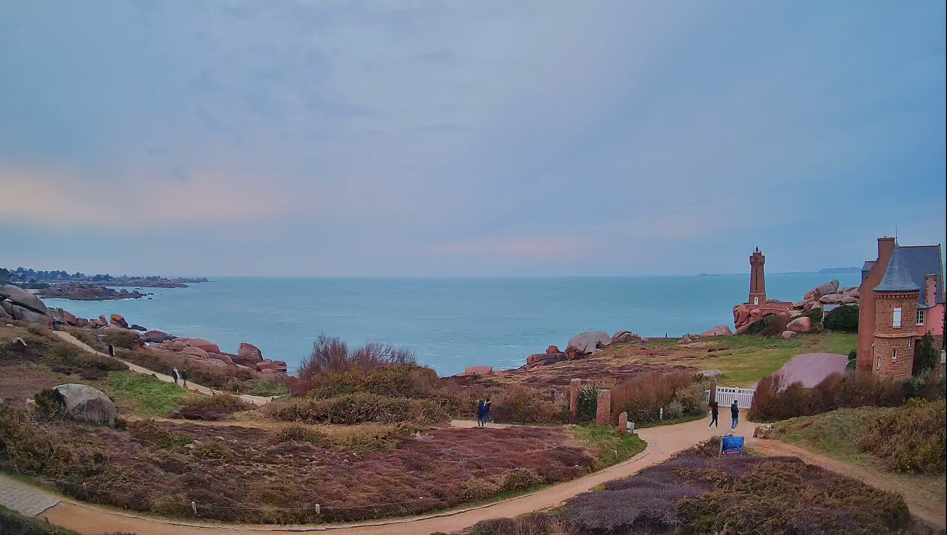 A coastal scene under a cloudy sky shows a path winding through low-lying vegetation toward a lighthouse and a stone building overlooking a calm sea.