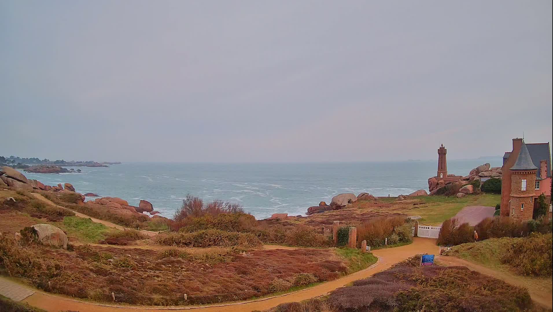 A coastal scene on an overcast day shows a rocky shoreline, a small lighthouse, a stone building, and a winding path through low-lying vegetation.