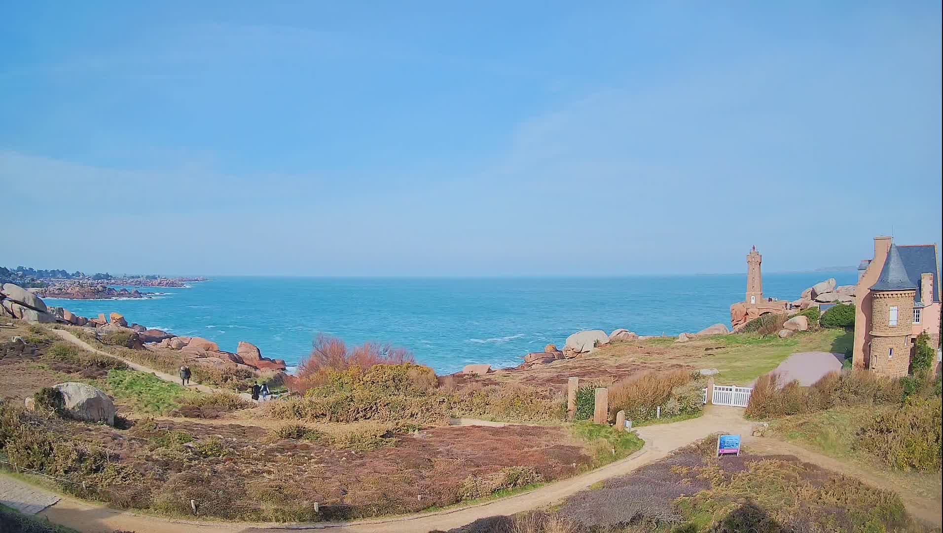 A coastal scene under a clear blue sky shows a lighthouse and stone building overlooking a turquoise ocean with rocky shoreline and a dirt path winding through low-lying vegetation.
