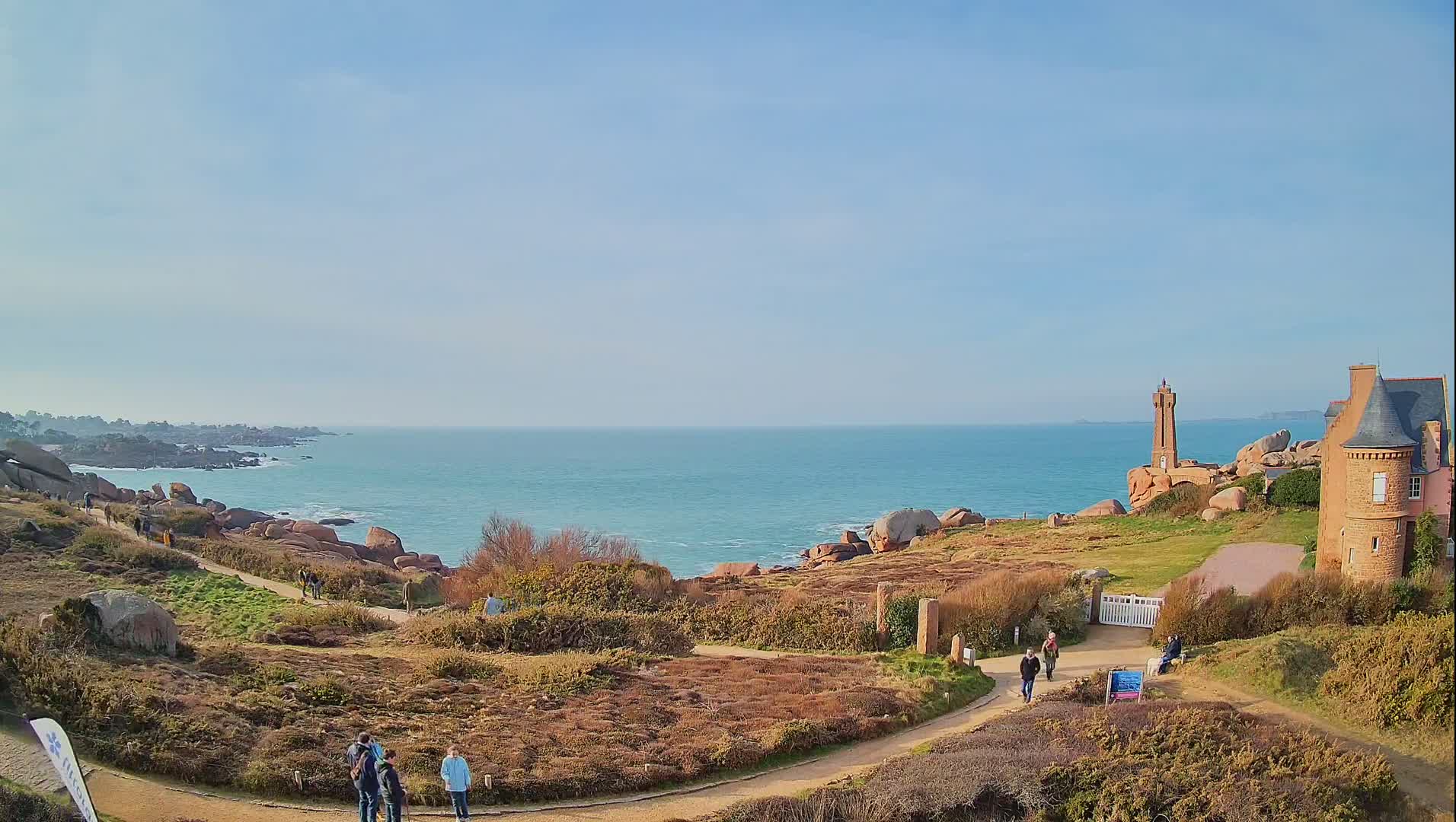 A coastal scene on a sunny day shows a stone path winding through low-lying vegetation toward a lighthouse and a stone building overlooking a calm, blue ocean dotted with pink rocks.