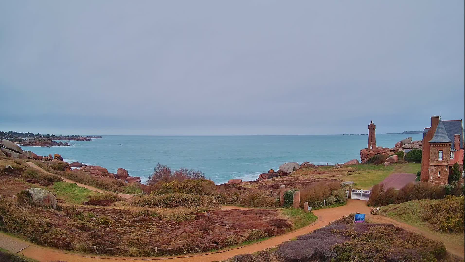 A coastal scene on an overcast day shows a red-brick building near a lighthouse overlooking a calm, teal ocean with pink granite rocks along the shore and winding paths through low, brown vegetation.