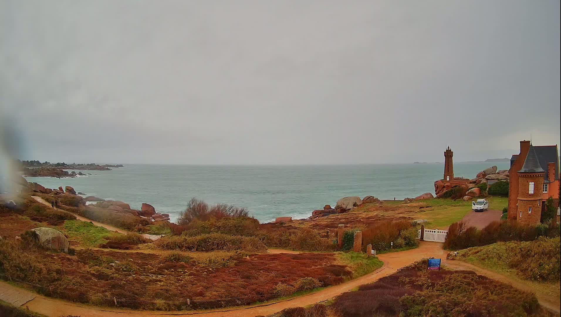 A coastal scene on an overcast day shows a path leading to a brick house and a small lighthouse situated on rocky terrain overlooking a calm sea.