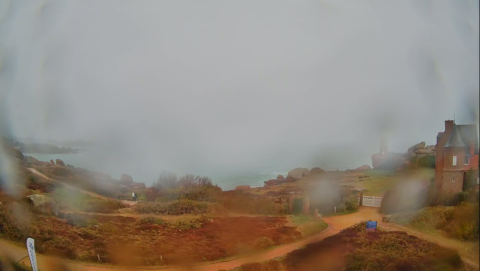 A foggy coastal scene shows a red brick house near a rocky shoreline with a path leading to a gate, viewed through rain-covered glass.