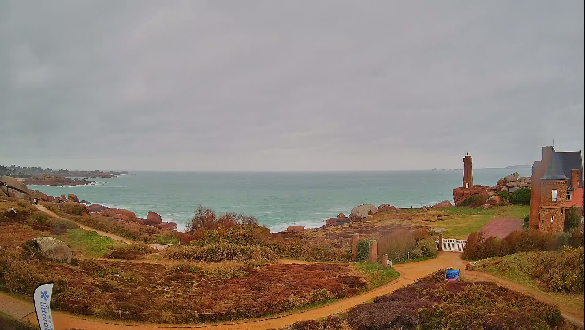 A coastal scene under an overcast sky shows a lighthouse and a stone house overlooking a rocky coastline and a calm sea.