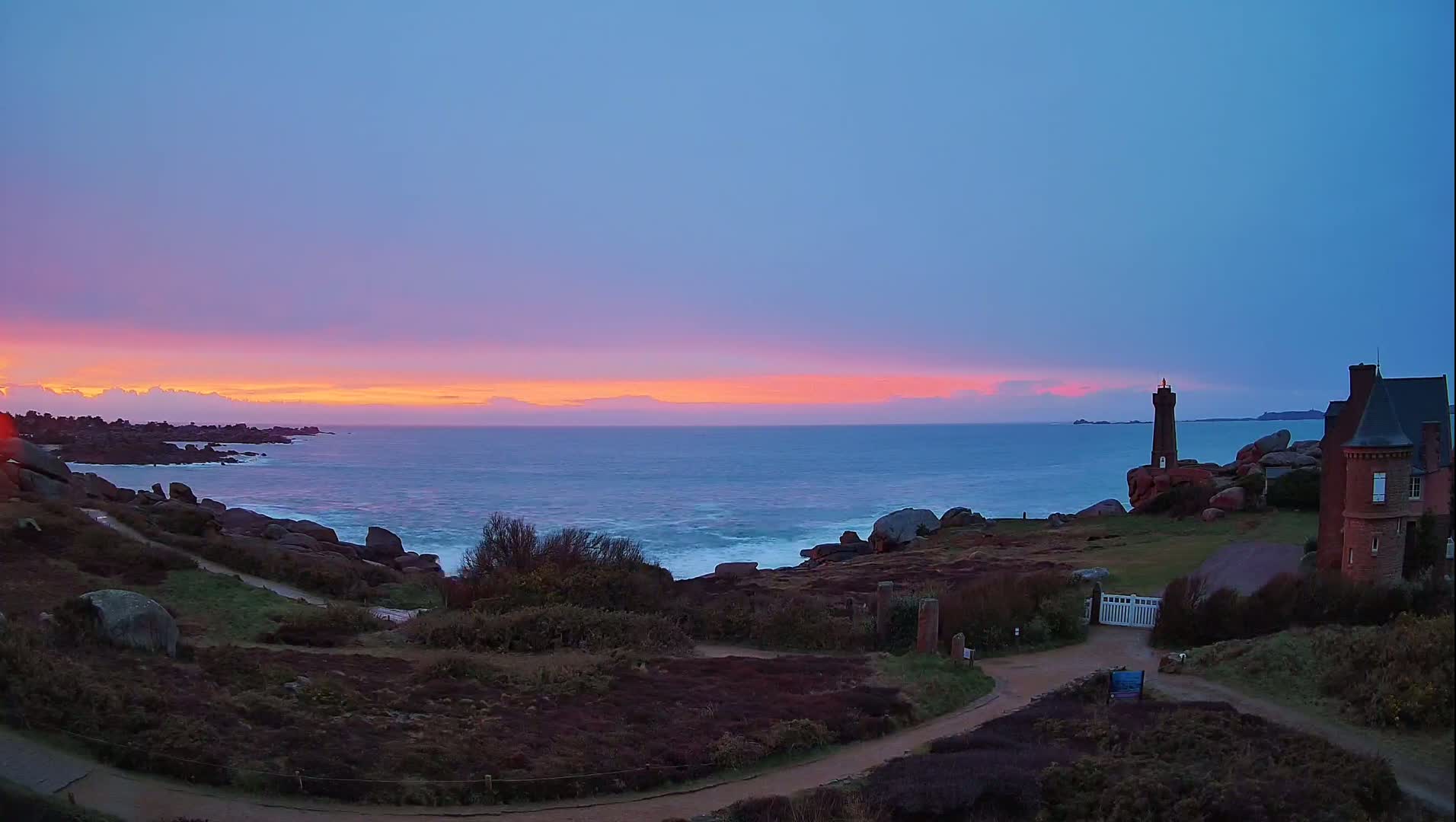 A rocky coastline at sunset features a lighthouse and a house overlooking a turbulent ocean under a colorful sky.