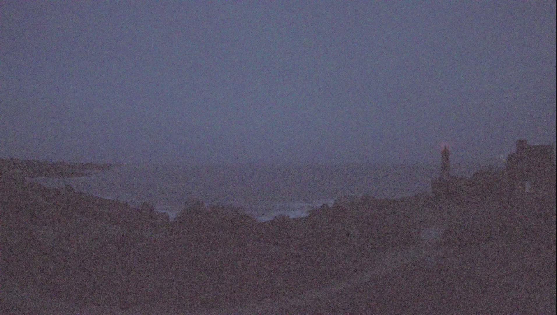 A hazy, dark-blue twilight sky overlooks a distant ocean and shoreline with a tall structure silhouetted against the horizon.