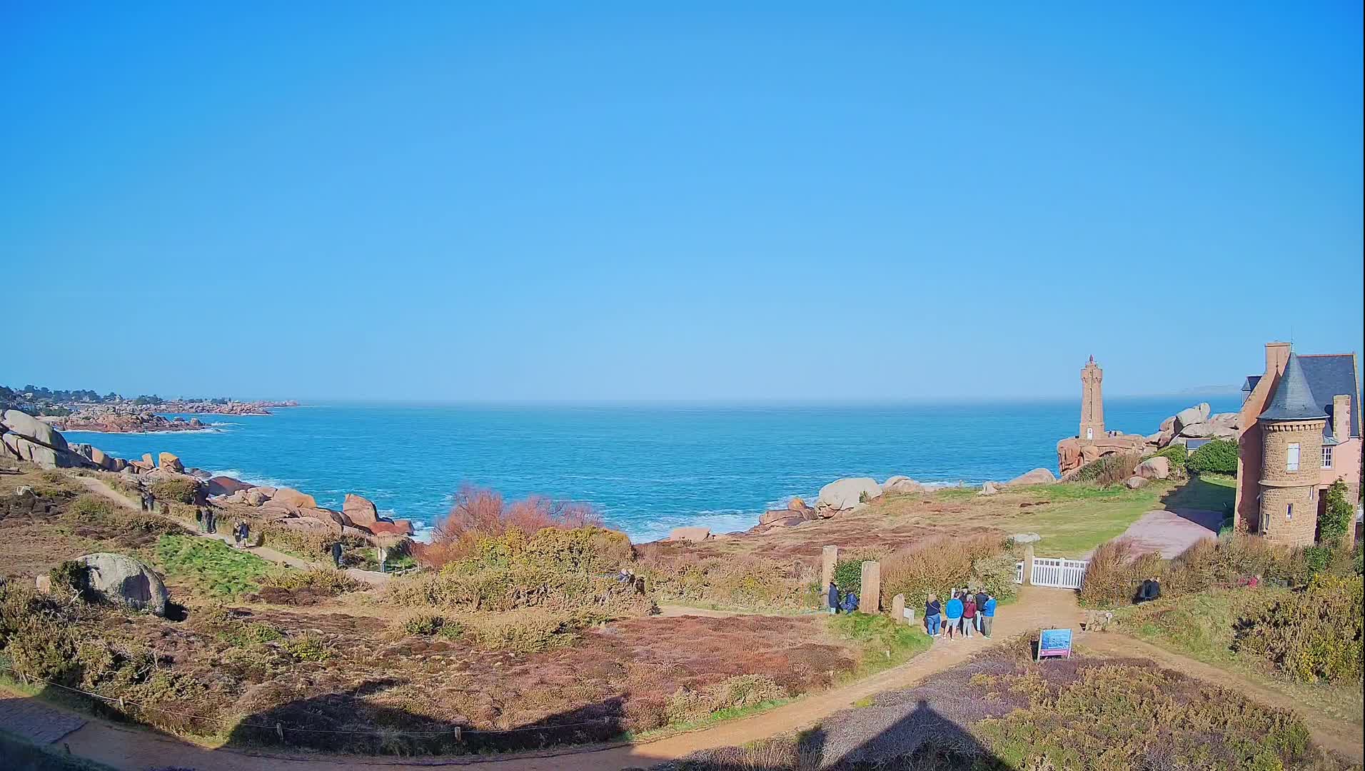 A coastal path winds through low-lying vegetation toward a lighthouse and a stone building overlooking a bright blue ocean on a clear, sunny day.