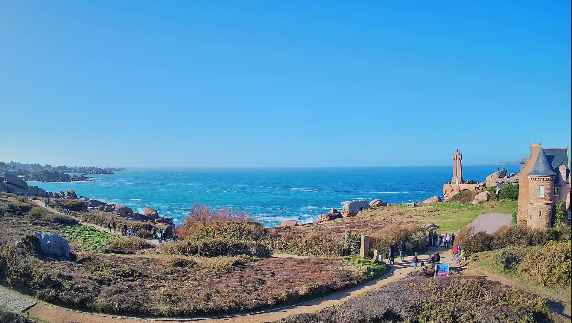 A coastal scene on a sunny day shows a rocky shoreline, a lighthouse, a stone building, and several people walking on paths overlooking a turquoise ocean with small waves.