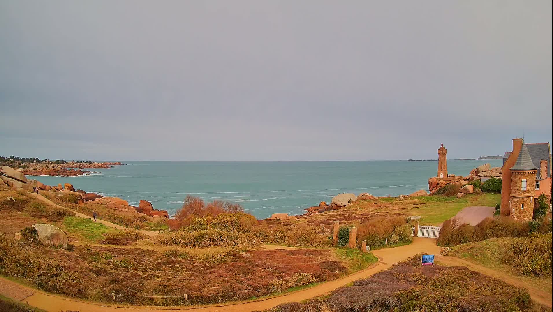 A coastal path winds through a rocky landscape overlooking a calm, teal sea under a cloudy sky, with a lighthouse and a stone building visible in the distance.