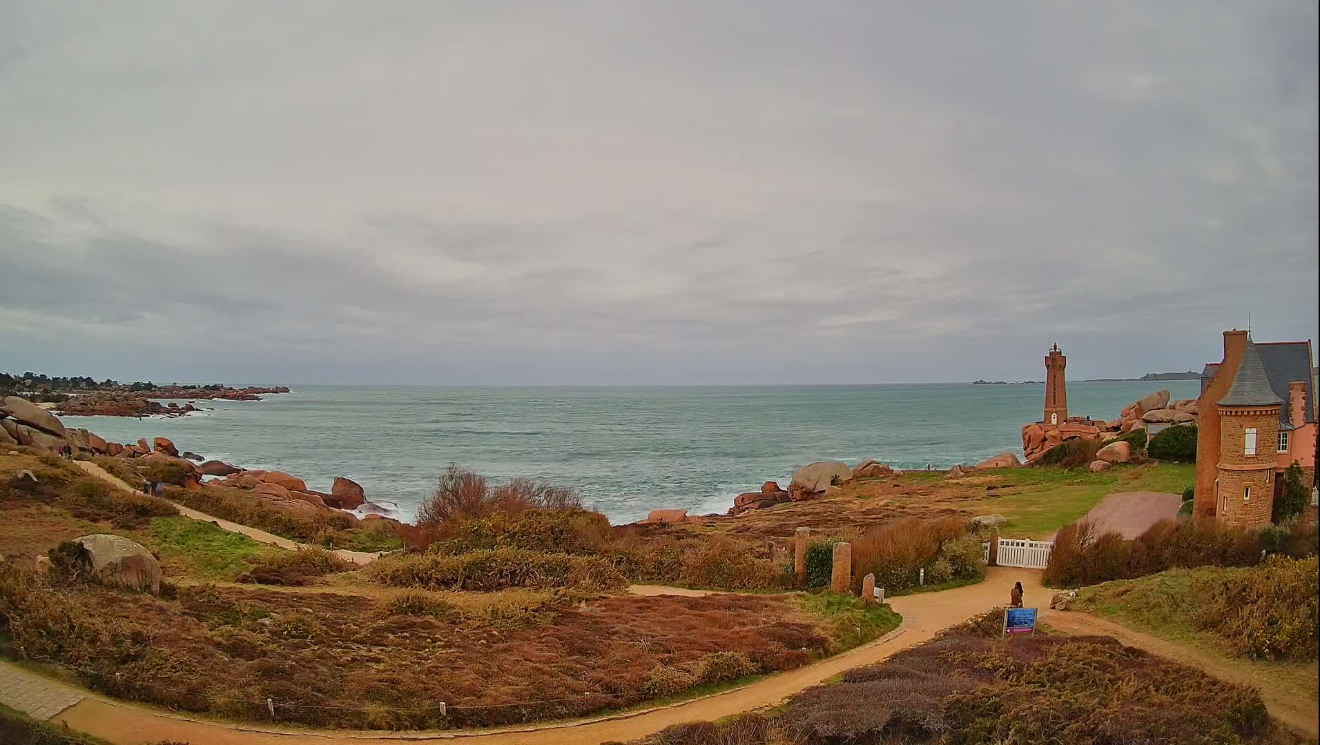 A coastal path winds through low-lying vegetation toward a lighthouse and a stone building overlooking a choppy sea under a cloudy sky.