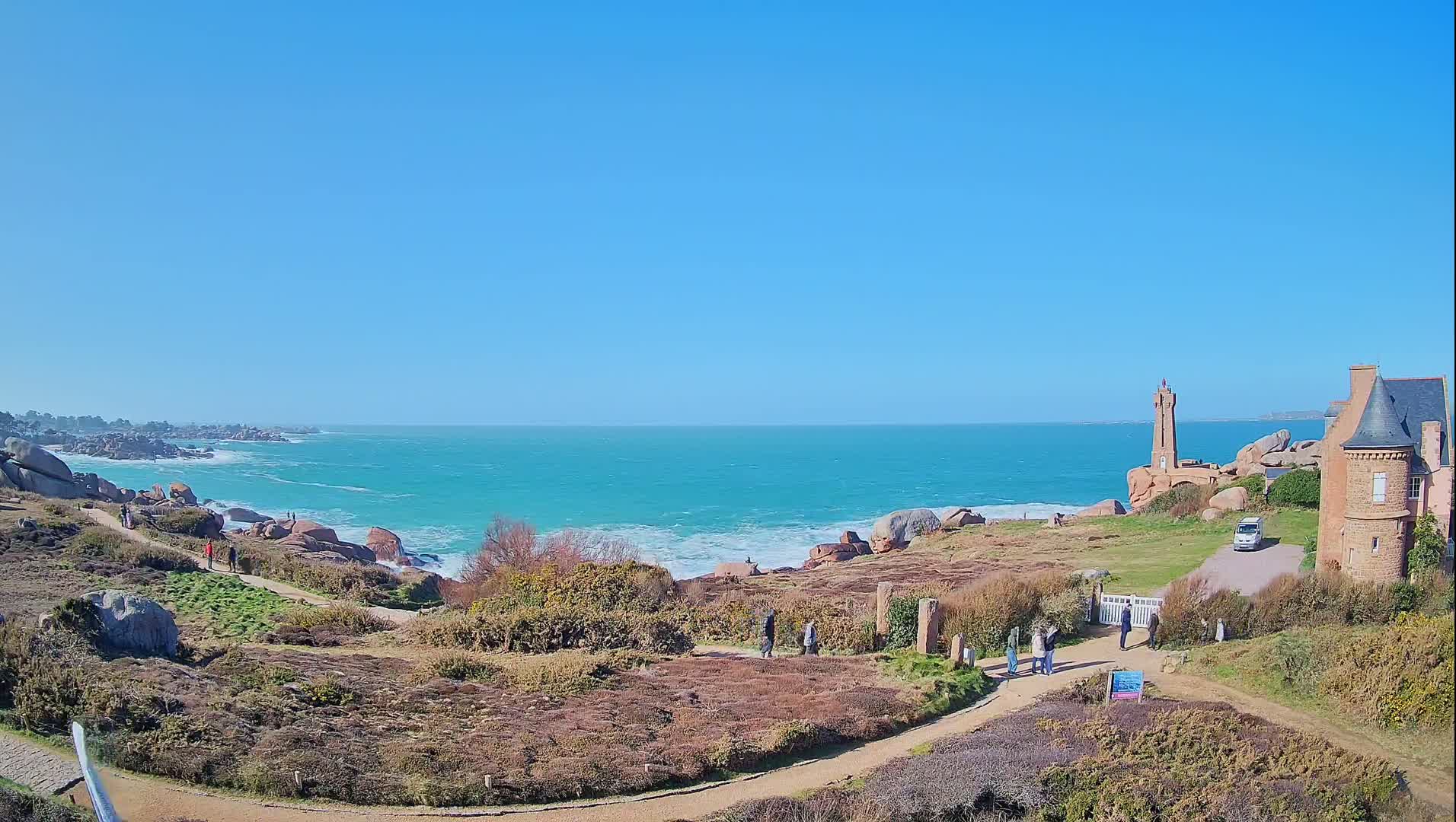 A coastal scene on a sunny day shows a rocky shoreline with a lighthouse and a stone building, winding paths, and a few people walking along the coast.