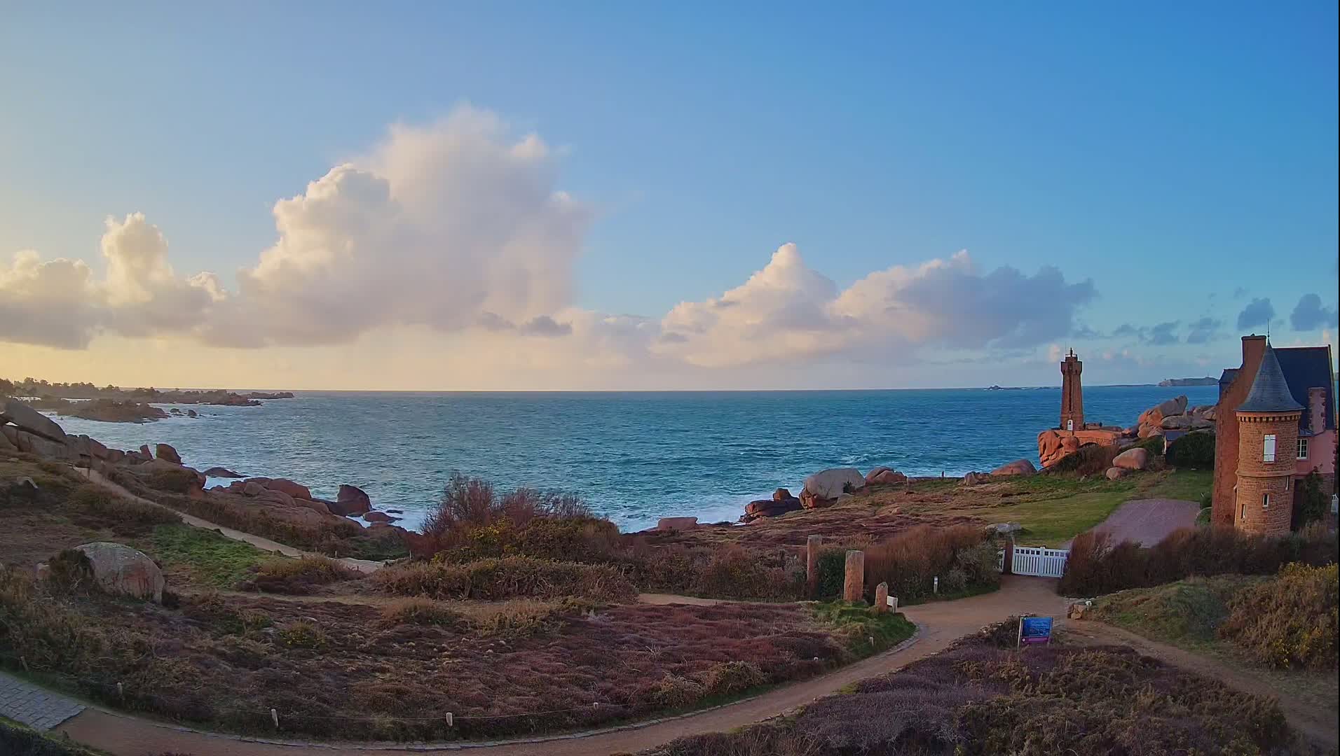 A rocky coastline with a lighthouse and a stone building overlooks a blue ocean under a partly cloudy sky.